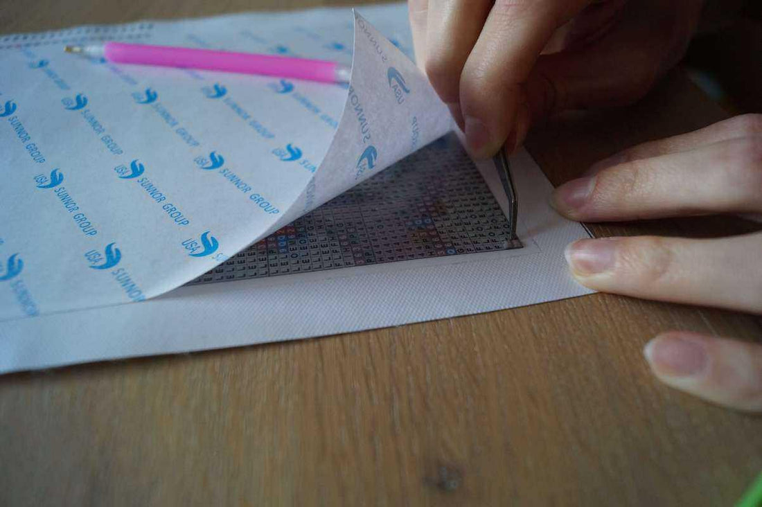Een hand tilt de beschermfolie van een diamond-paintingcanvas